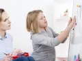 Terapeutaka z piórem w dłoni obserwuje dziewczynkę przyczepiającą rysunki do tablicy, analizując jej słabe strony.