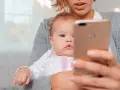 Mama trzyma na rękach niemowlę, które ma około 20 miesięczne. Dziecko patrzy na telefon.