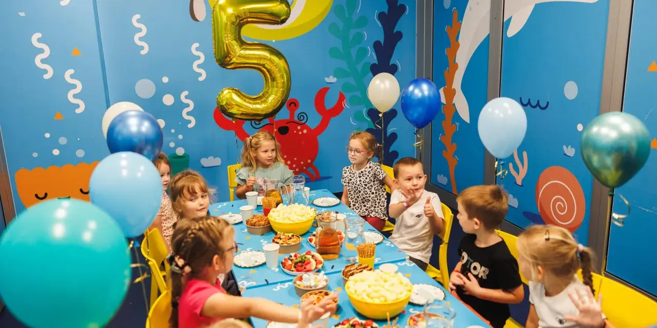 Dzieci świętują 5 urodziny przy stole pełnym przekąsek. Złoty balon w kształcie cyfry 5 unosi się nad stołem.
