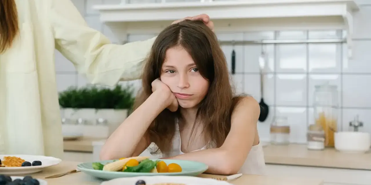 Smutna dziewczynka przy stole, oparta o dłoń, wyraźnie pokazuje, że dziecko nie chce jeść. Mama głaszcze ją po głowie.