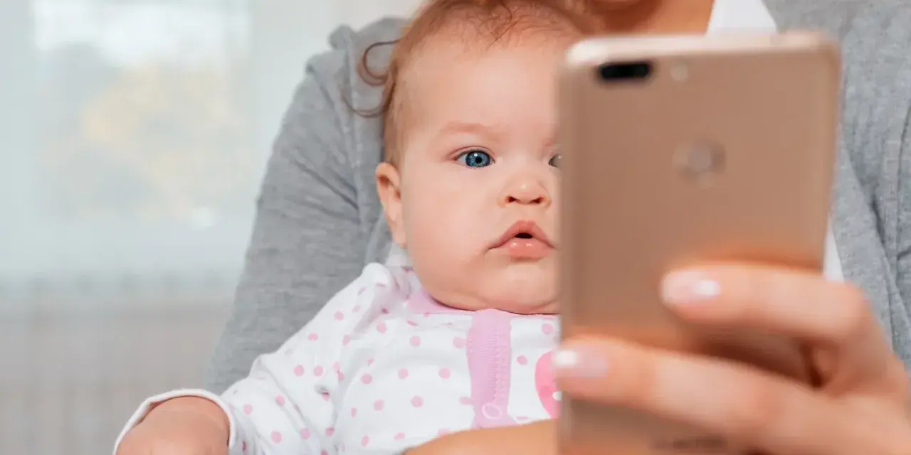Mama trzyma na rękach niemowlę, które ma około 20 miesięczne. Dziecko patrzy na telefon.