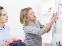 Terapeutaka z piórem w dłoni obserwuje dziewczynkę przyczepiającą rysunki do tablicy, analizując jej słabe strony.