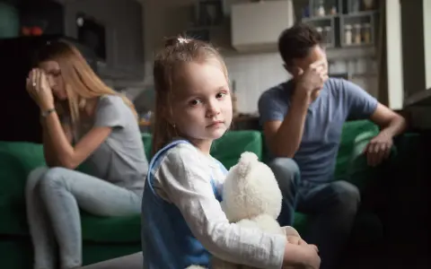 Smutna dziewczynka z misiem w objęciach, podczas gdy rodzice w tle przeżywają trudne chwile. Temat: rozwód a dzieci.