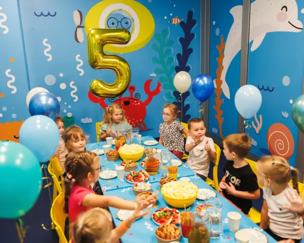 Dzieci świętują 5 urodziny przy stole pełnym przekąsek. Złoty balon w kształcie cyfry 5 unosi się nad stołem.