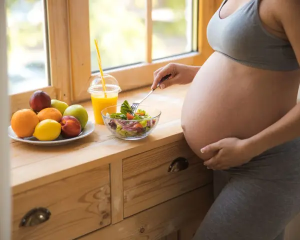 Kobieta w 39 tygodniu ciąży je sałatkę. Obok stoi talerz z owocami i sok pomarańczowy.