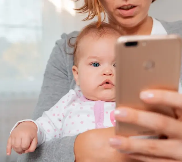 Mama trzyma na rękach niemowlę, które ma około 20 miesięczne. Dziecko patrzy na telefon.