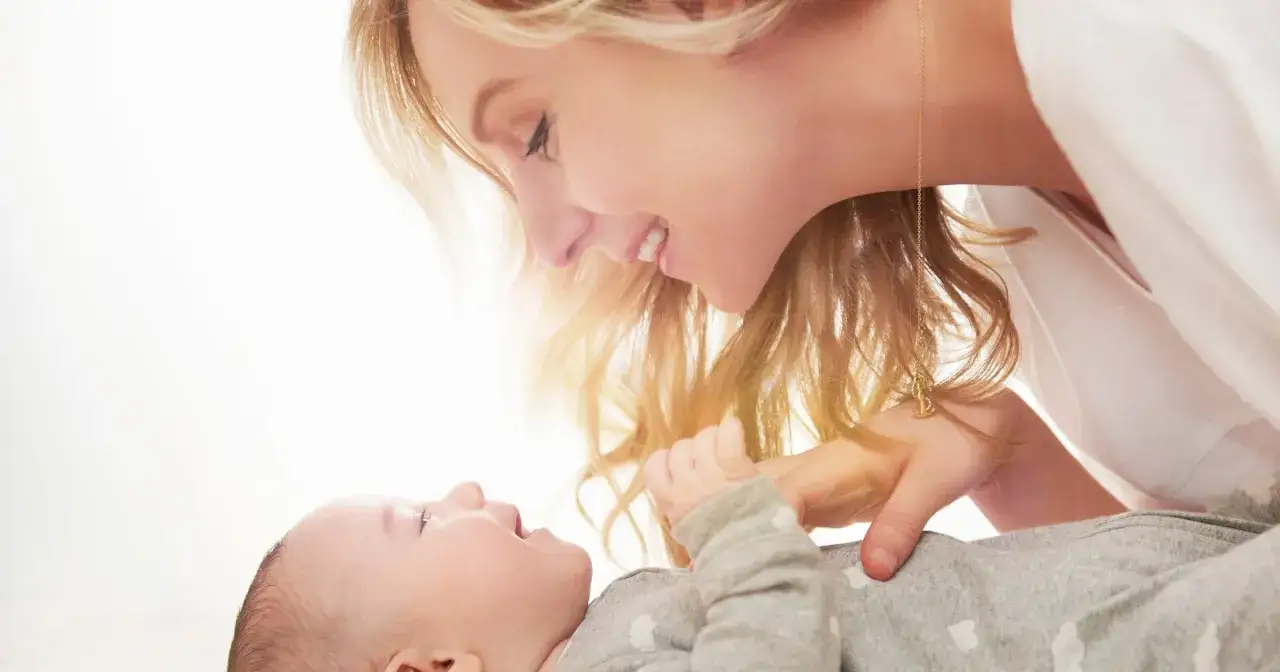 Uśmiechnięta mama pochyla się nad dzieckiem, które ma czkawkę u noworodka. Delikatne światło otula ich twarze.